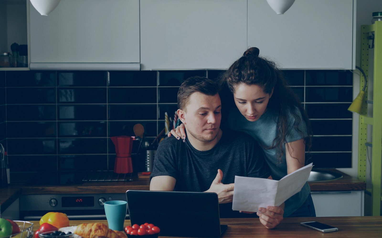 Para przy stole w kuchni czyta pisma z banku, przed nimi laptop